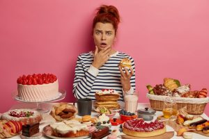 Indignant red haired woman in striped jumper shocked how much calories she ate for day, holds sweet dessert, has no diet, isolated over rosy wall, eats sweet food. Bakery, confectionery concept
